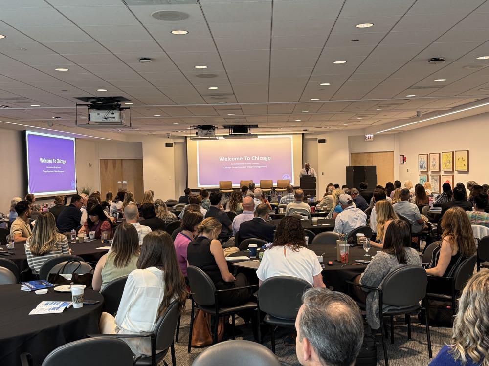 Large conference room full of attendees watching a stage presentation with two projection screens reading 'Welcome To Chicago'