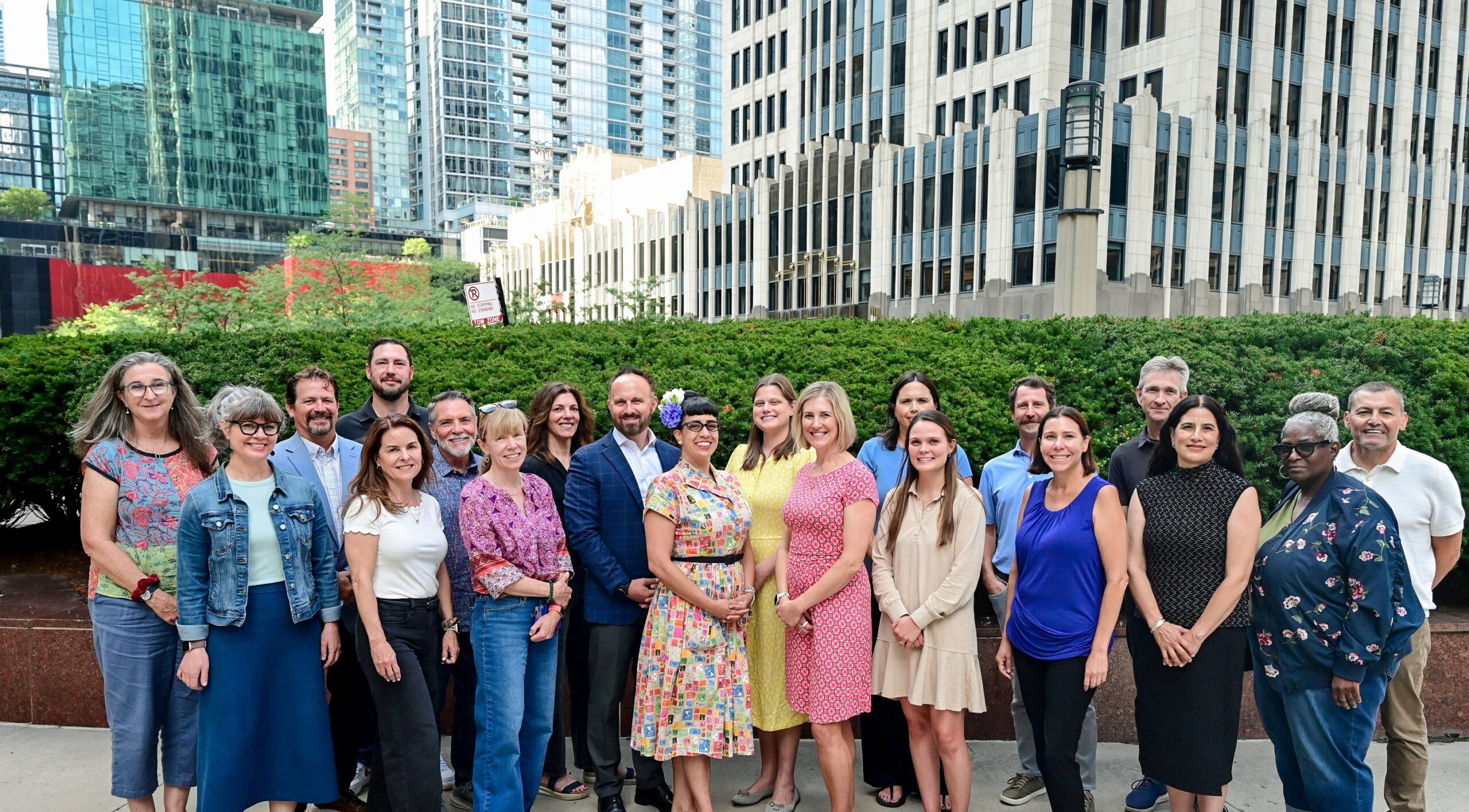 Diverse group of about 20 adults posing together outdoors in front of modern skyscrapers and a hedge backdrop.