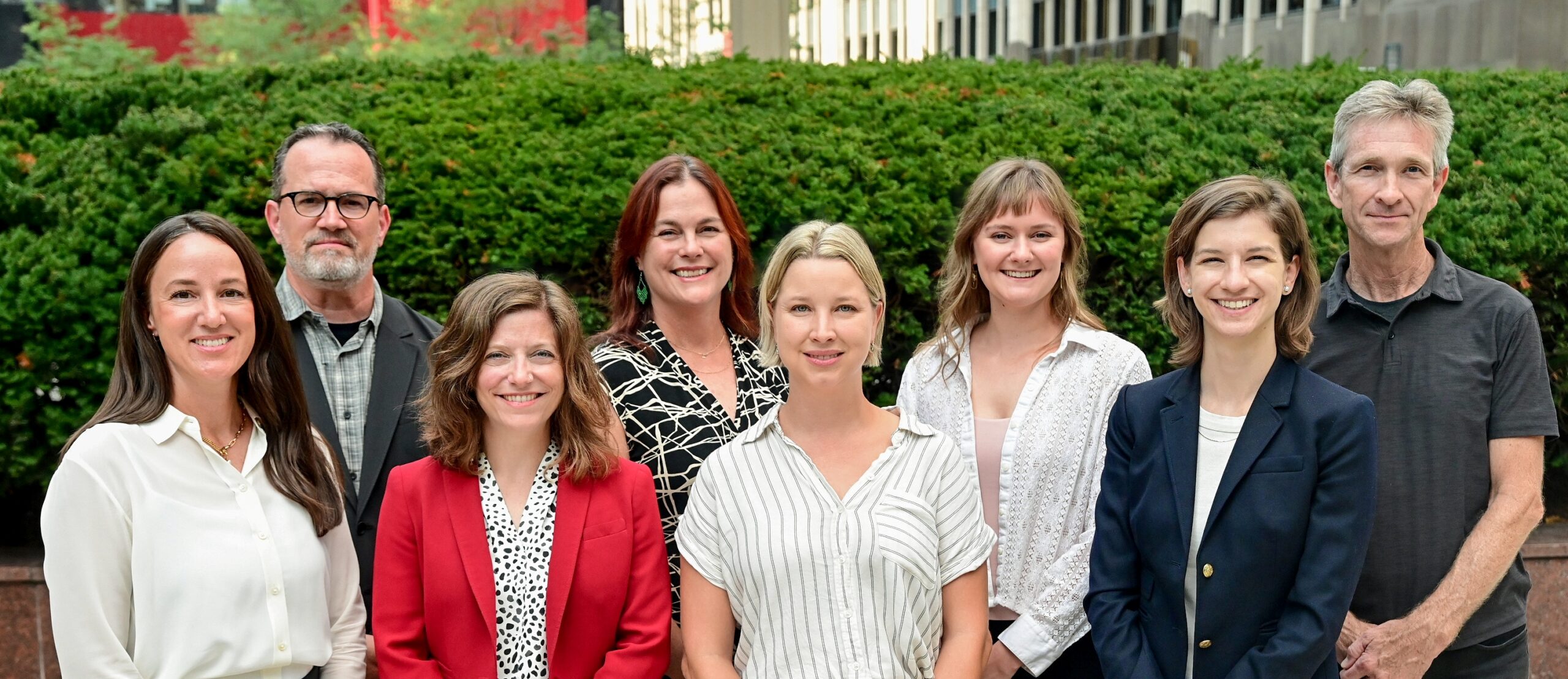 Eight adults posing outdoors in front of a hedge, smiling at the camera.