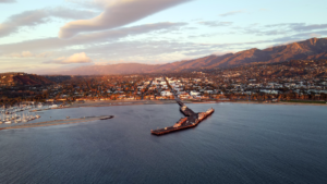 Aerial view of a coastal town at sunset with a long pier extending into the harbor and mountains in the distance.