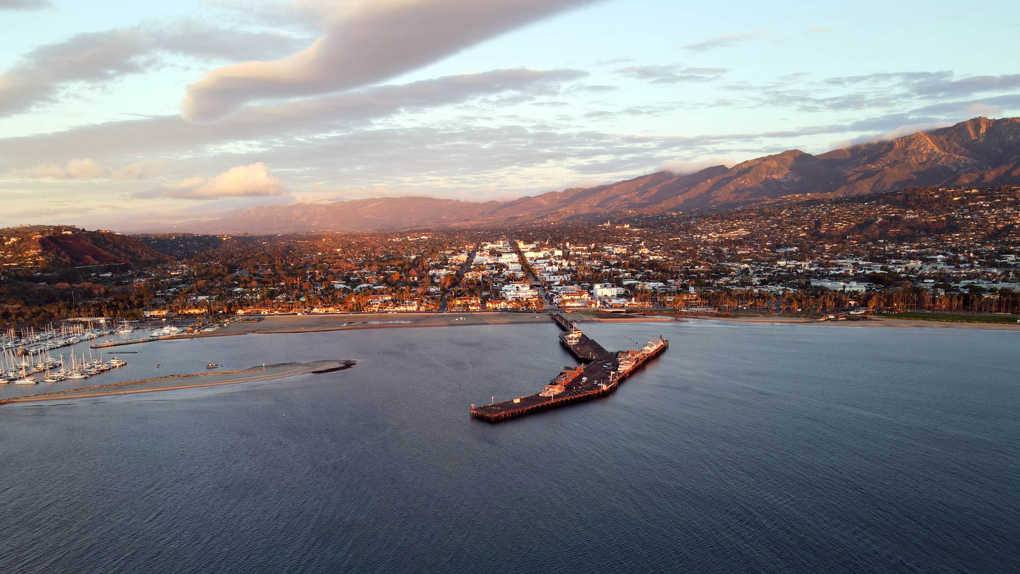 Aerial view of a coastal town at sunset with a long pier extending into the harbor and mountains in the distance.