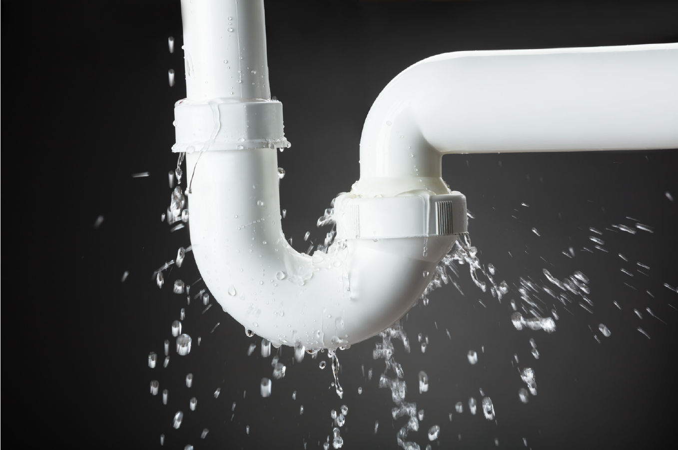 Close-up of a white PVC pipe elbow with water droplets spraying against a dark background, emphasizing plumbing leaks.